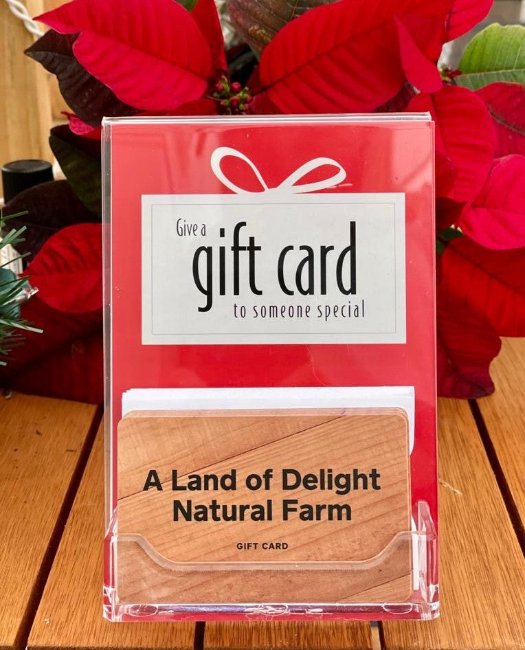 Small display holding Woodgrain Land of Delight Natural Farm Gift Card in front of a poinsietta plant on a wooden table